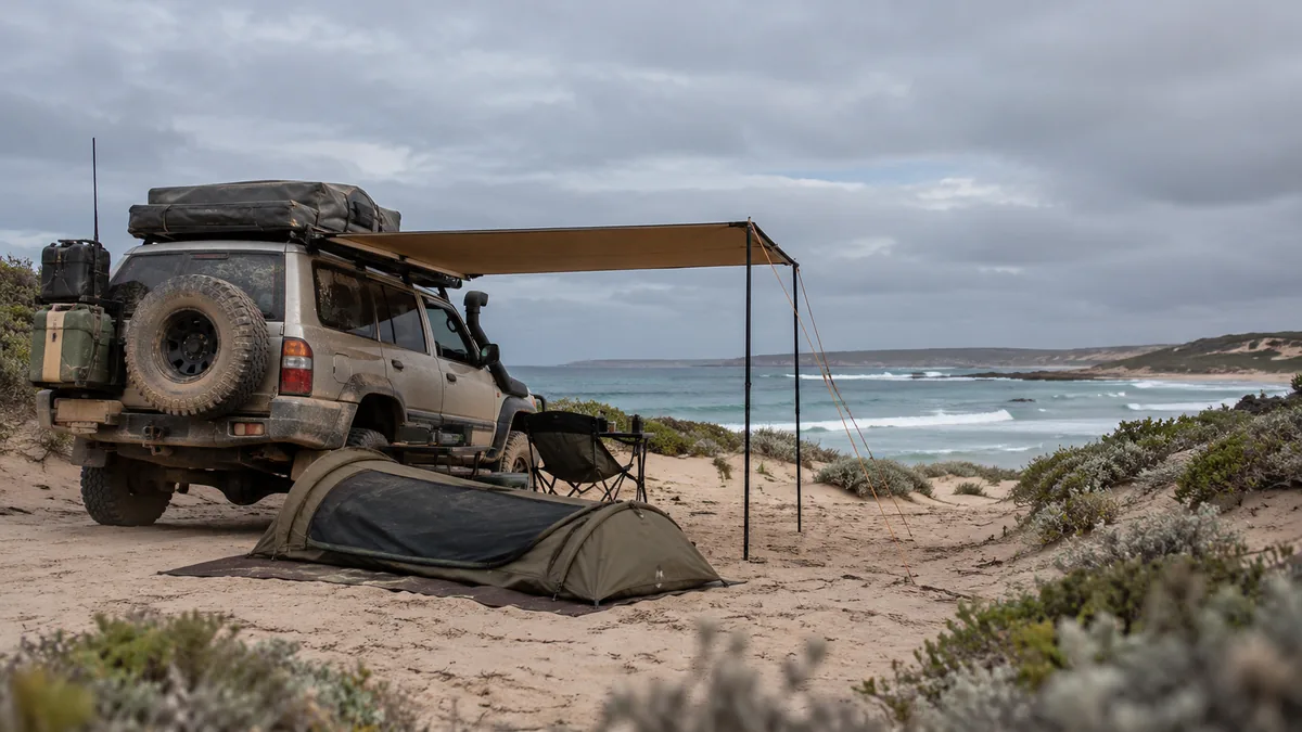Dusty 4WD with awning and swag set up on sand behind dunes at an Australian beach